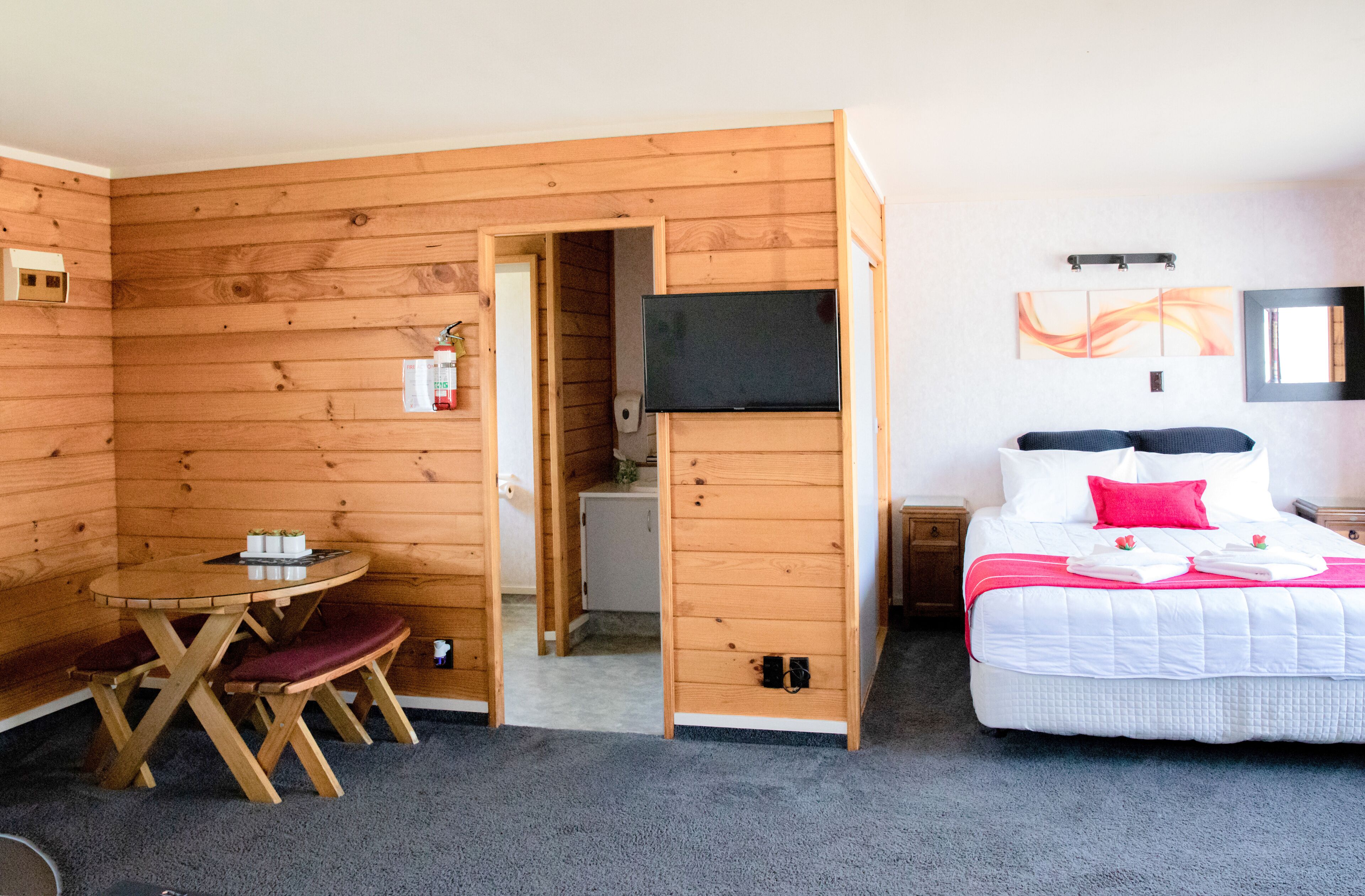 A wooden cabin-style room with a bed, a TV, a small table with chairs, and a door leading to a bathroom.
