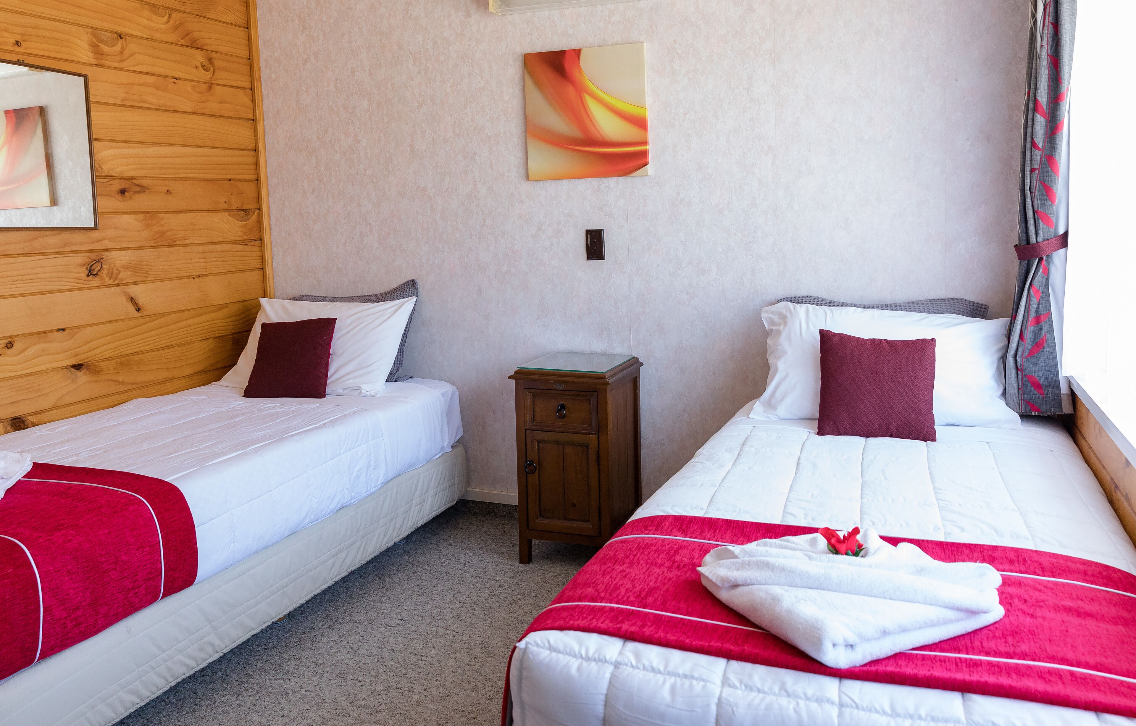 Habitación de hotel con dos camas, cada una con ropa de cama blanca y detalles rojos, una mesita de noche de madera y un cuadro en la pared.