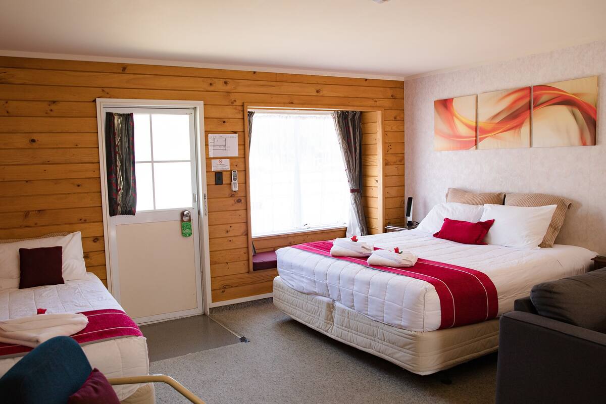 A hotel room with two beds, wooden walls, a window with curtains, a painting on the wall, and a telephone on the nightstand.