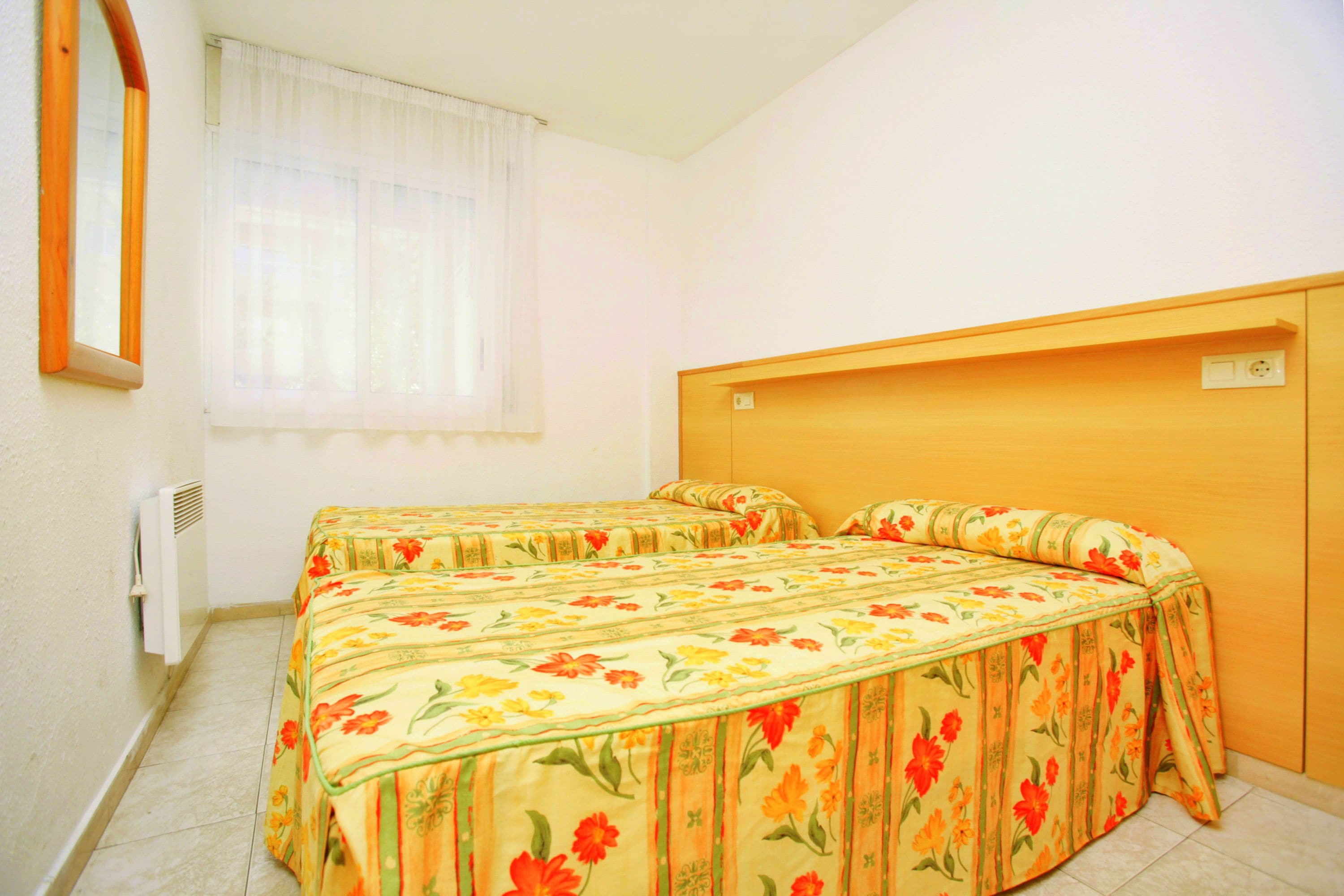 A room with two beds, a window with sheer curtains, a radiator, and a wooden headboard.