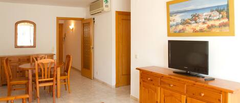 Una sala de estar con una mesa de comedor de madera, sillas, un banco, un televisor sobre un mueble de madera y un cuadro en la pared.