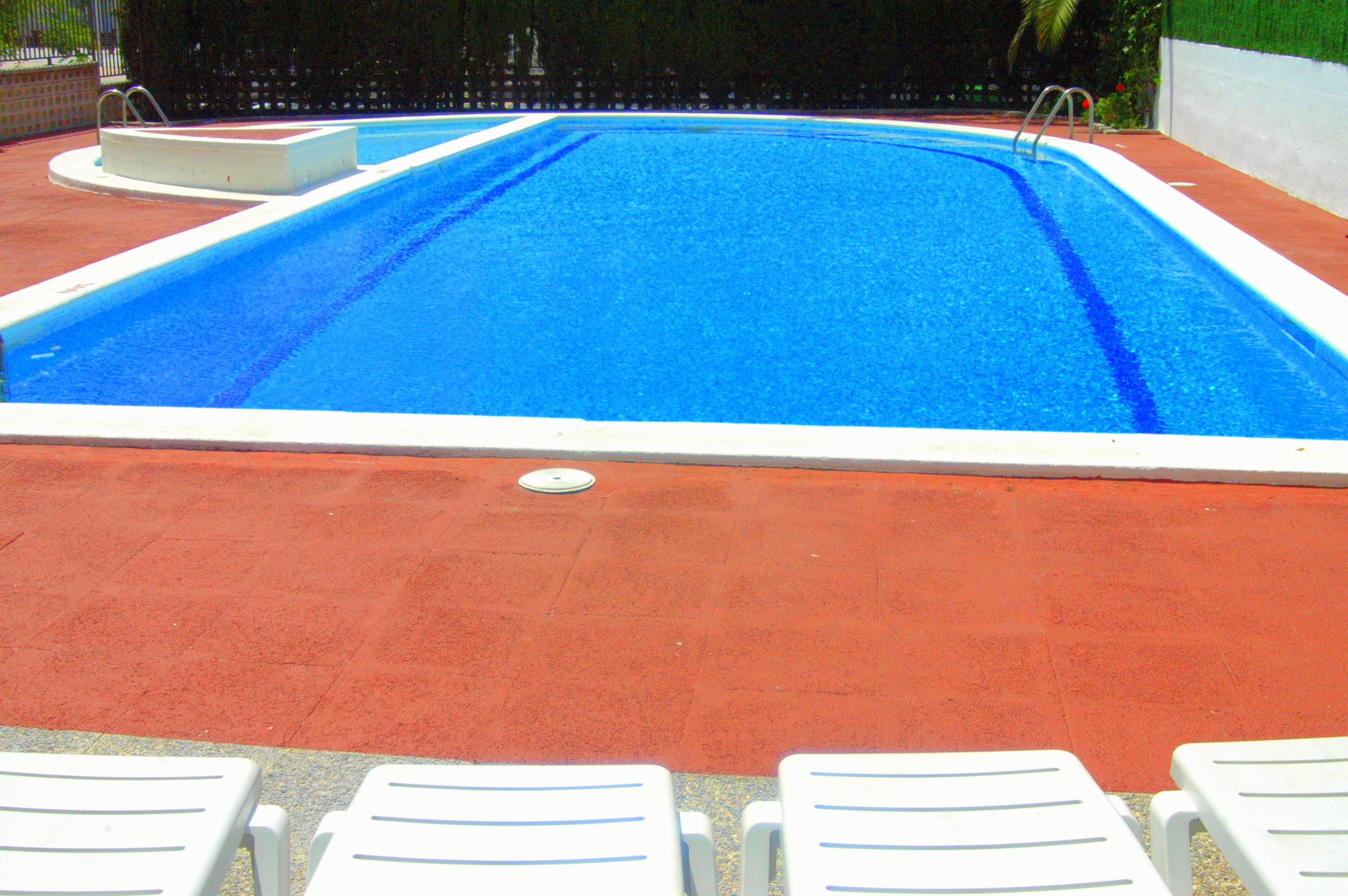 Una piscina rectangular con borde blanco y parte interior azul. Cuatro sillas reclinables blancas están alineadas junto a la piscina.