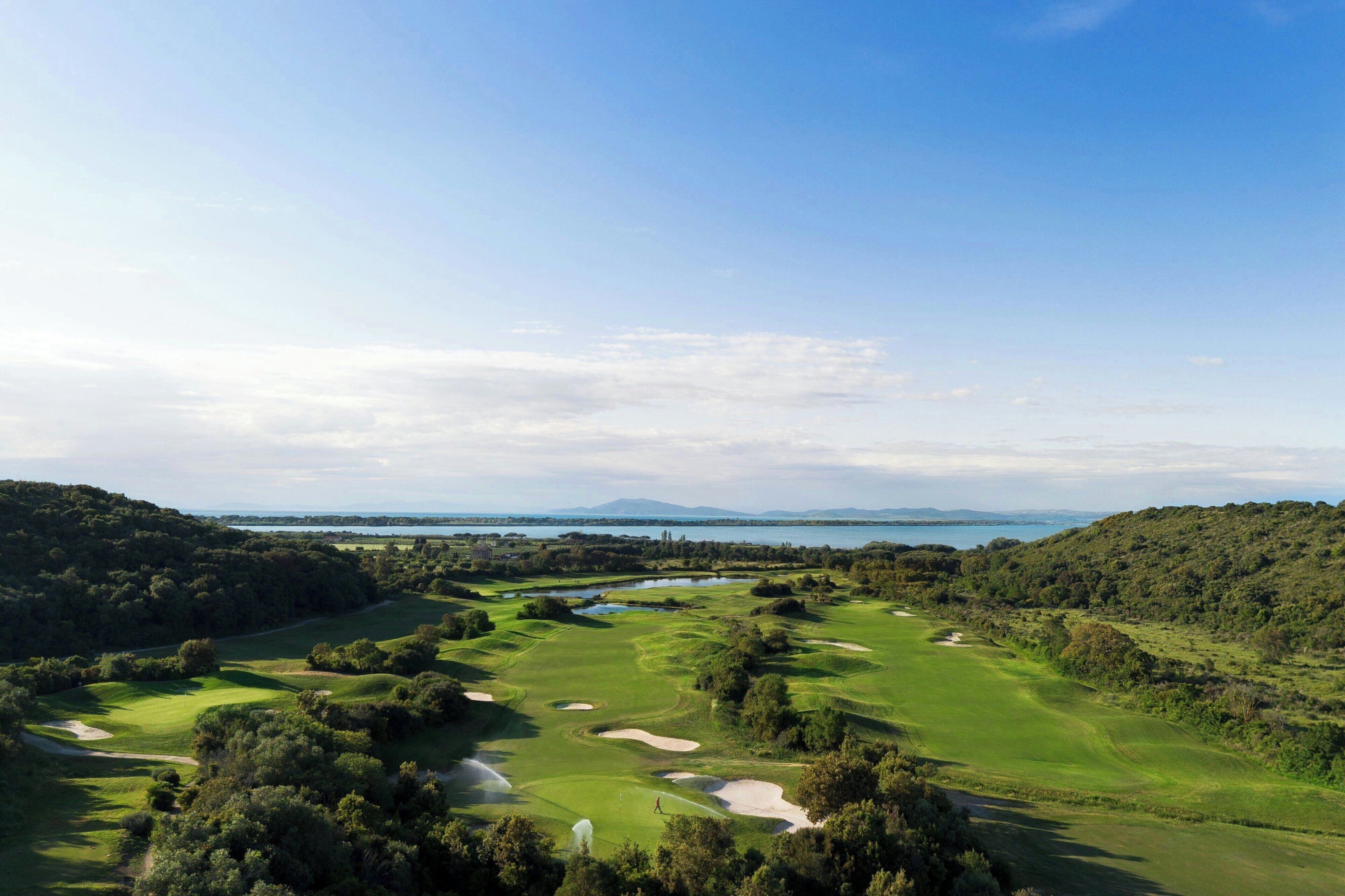 A golf course with a water body in the background, surrounded by greenery.