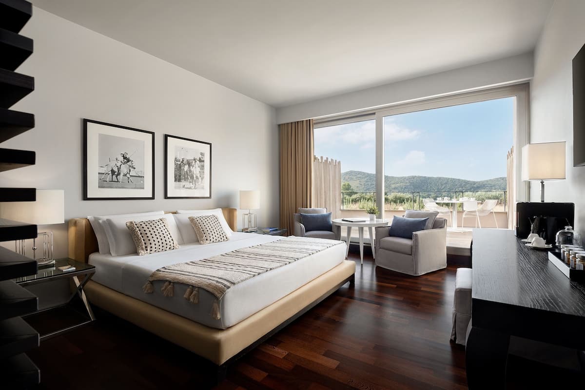 A modern hotel room with a large bed, a desk, a chair, and a view of the outdoors.