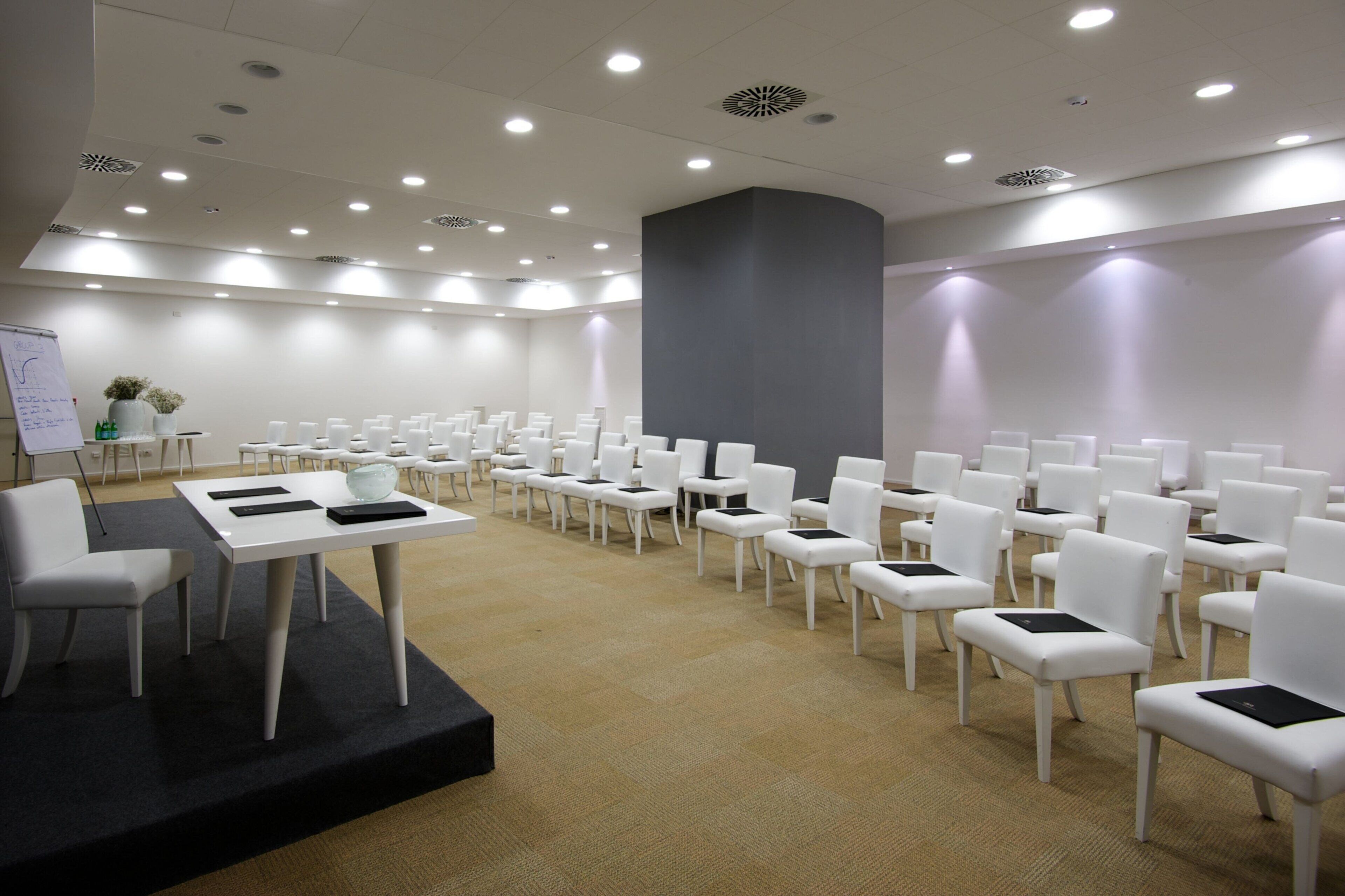 Una sala conferenze con file di sedie bianche, un tavolo bianco con tablet neri e una lavagna con appunti.