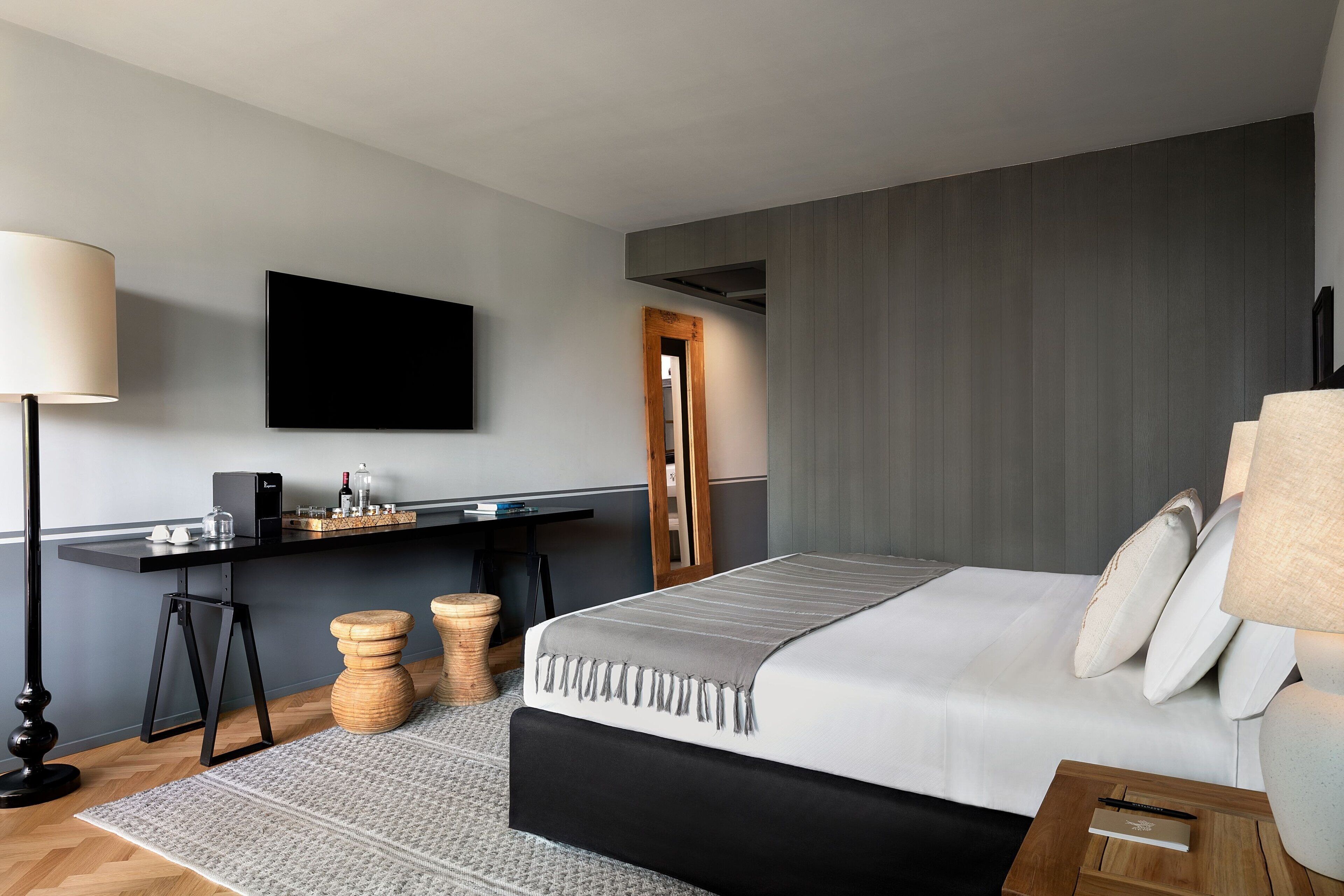 A modern hotel room with a large bed, a desk with a flat-screen TV, and a wooden side table.