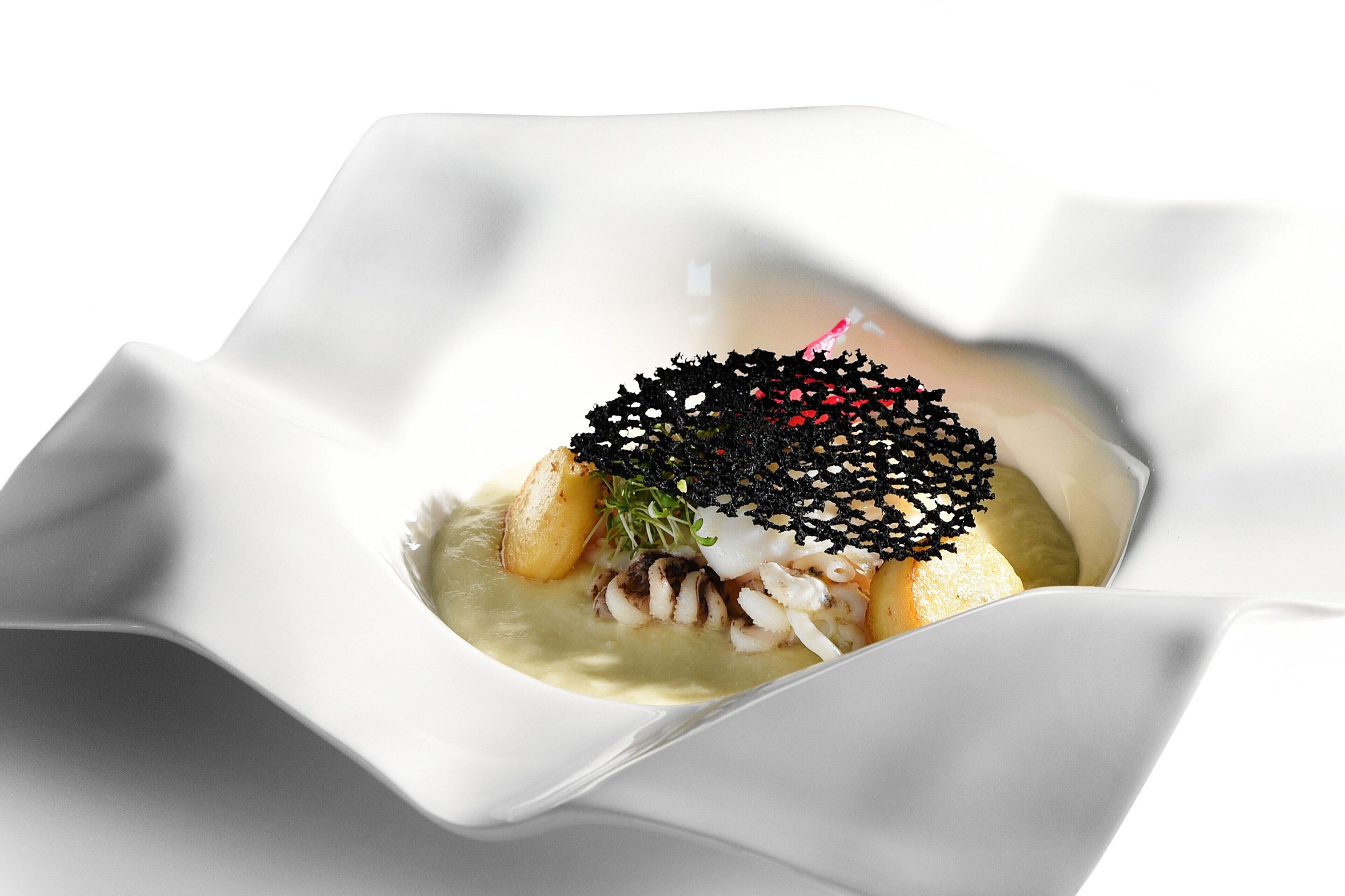 A white, uniquely shaped dessert dish with a central mound of food, surrounded by a creamy sauce and garnished with green herbs and black edible beads.