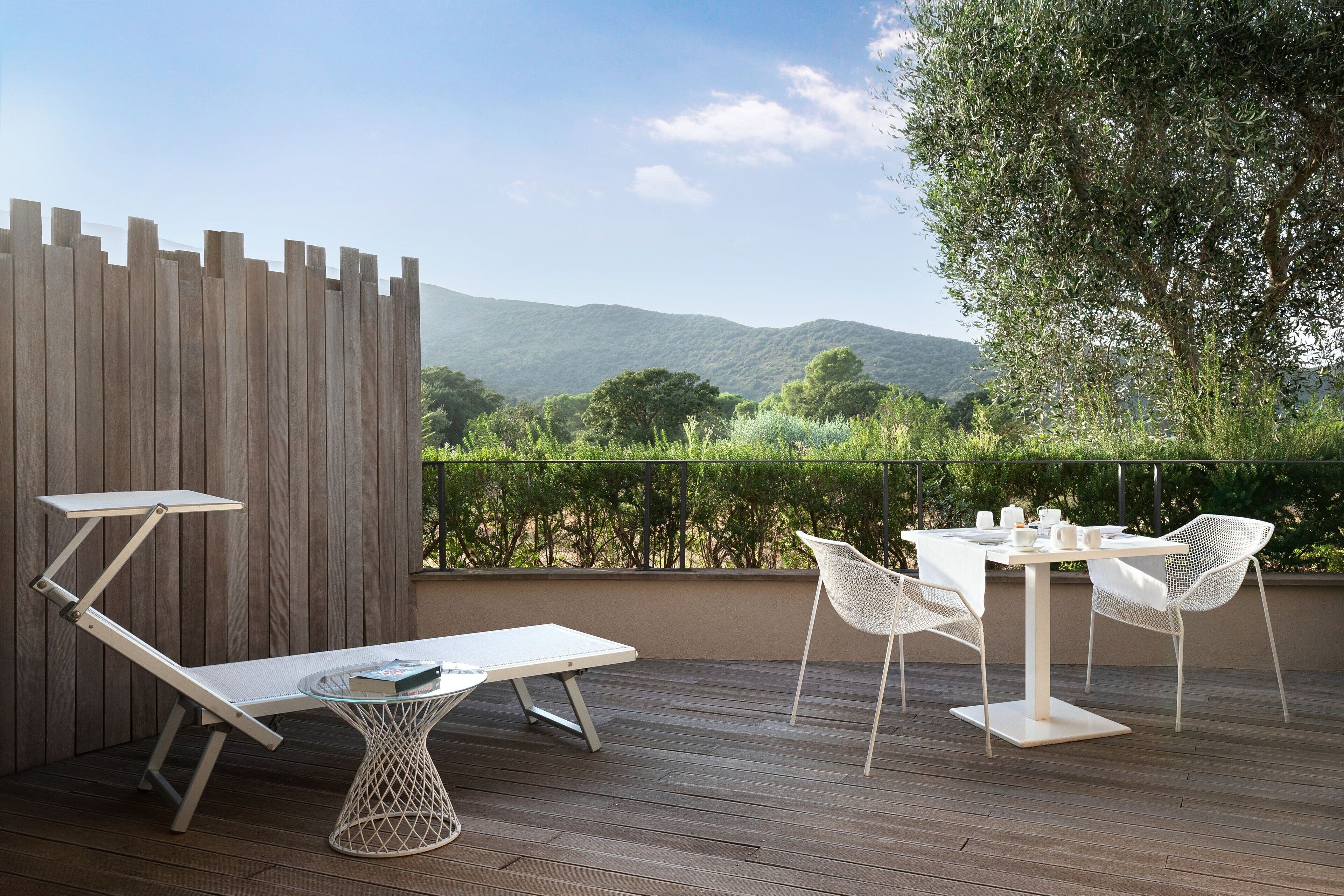 A wooden deck with a white table and chairs, a white lounge chair, and a view of green hills and trees.
