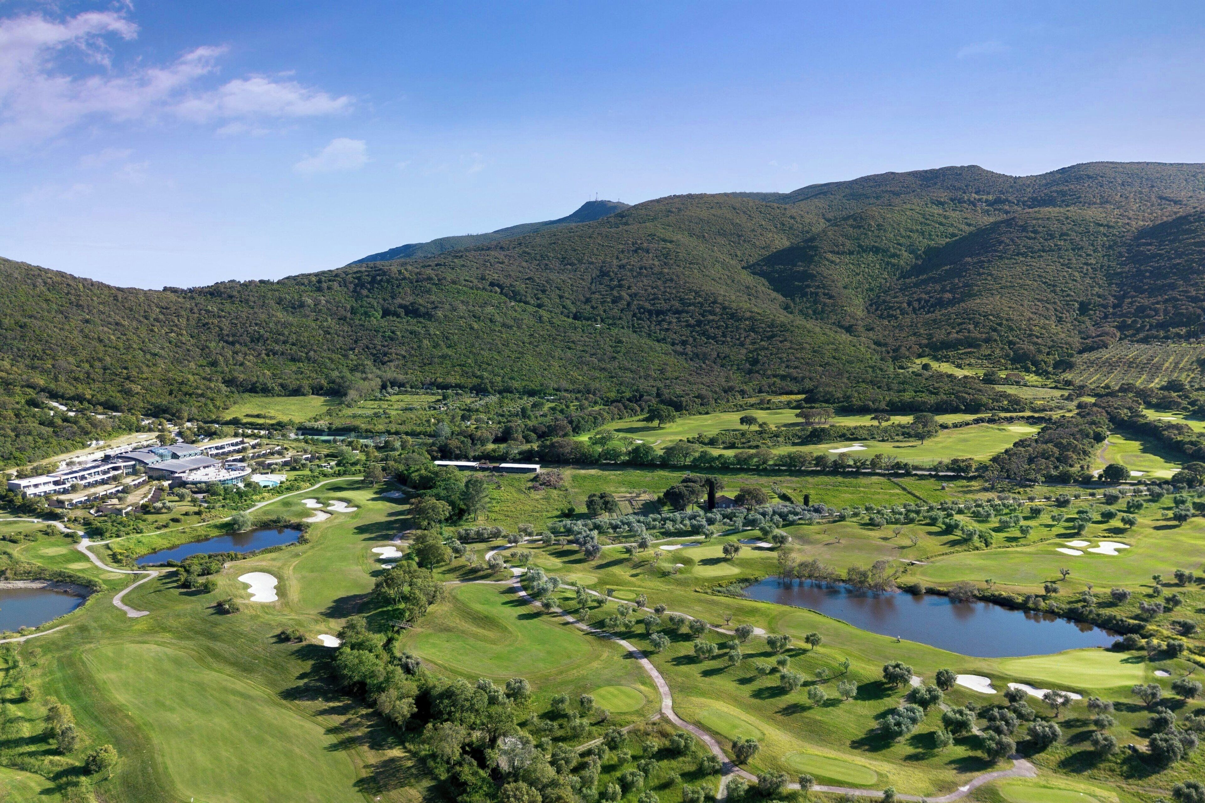 A golf course with multiple ponds, surrounded by lush green hills and a large building complex.