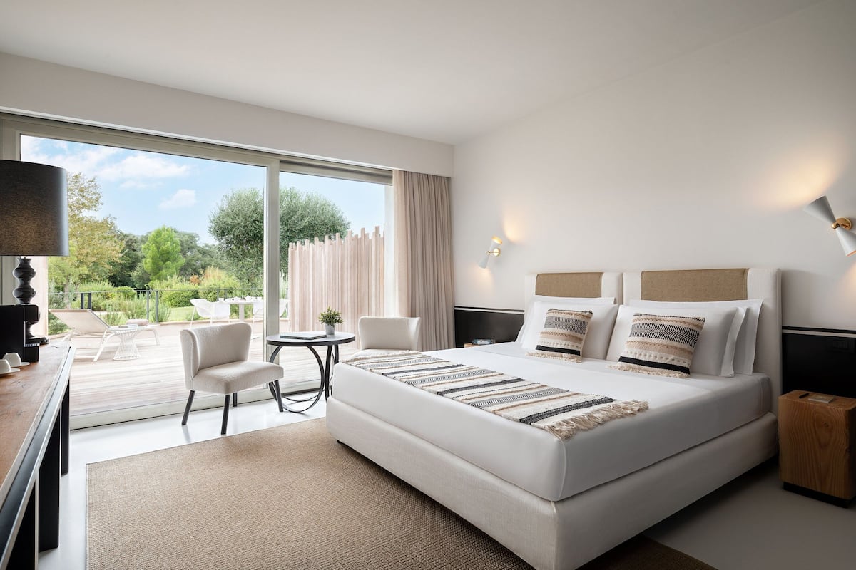 A modern hotel room with a large bed, two bedside tables, a desk, and a chair. The room has a large window offering a view of the outdoors.
