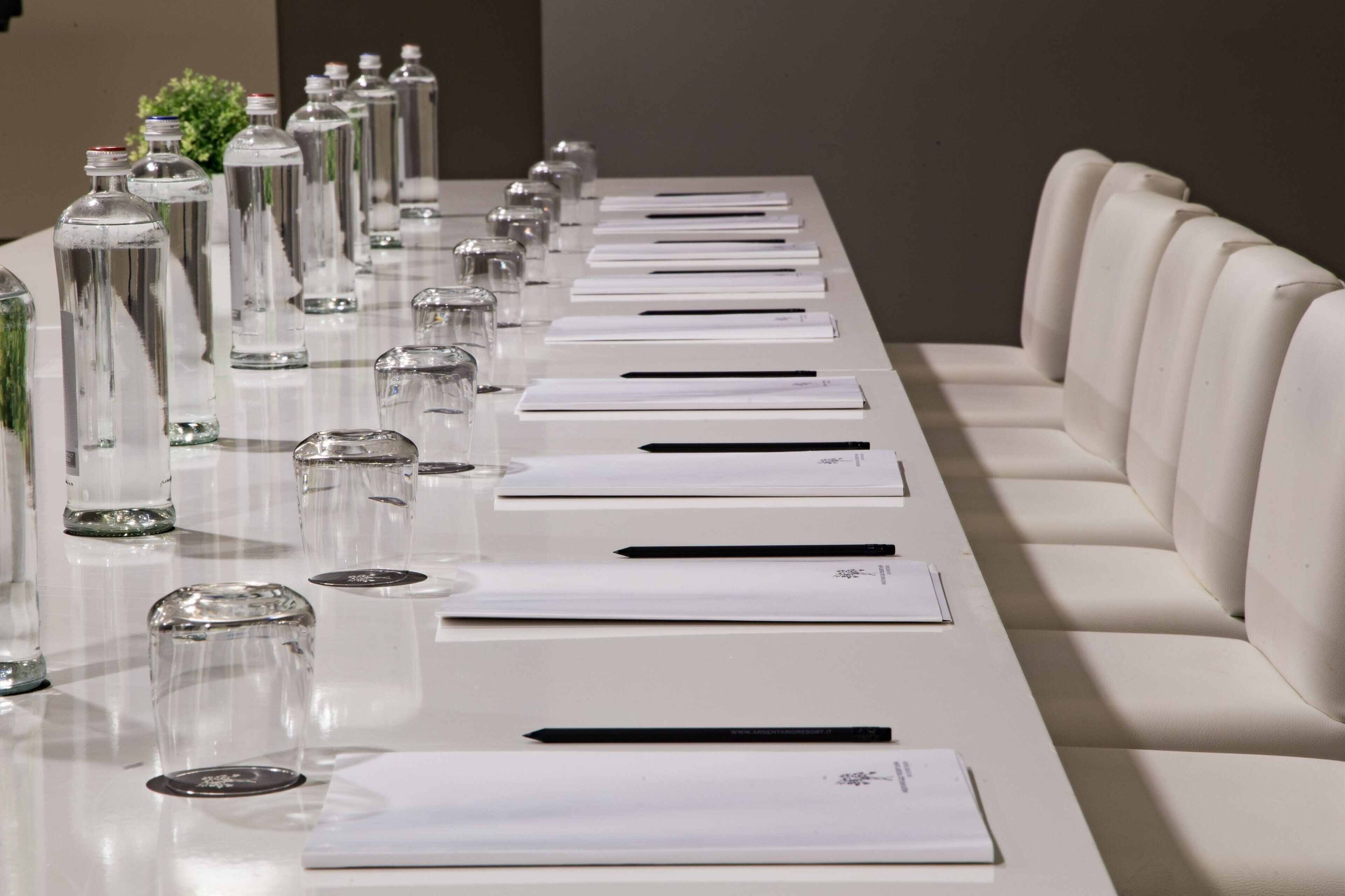 A long, white conference table with clear glass bottles, white notepads, and pens.