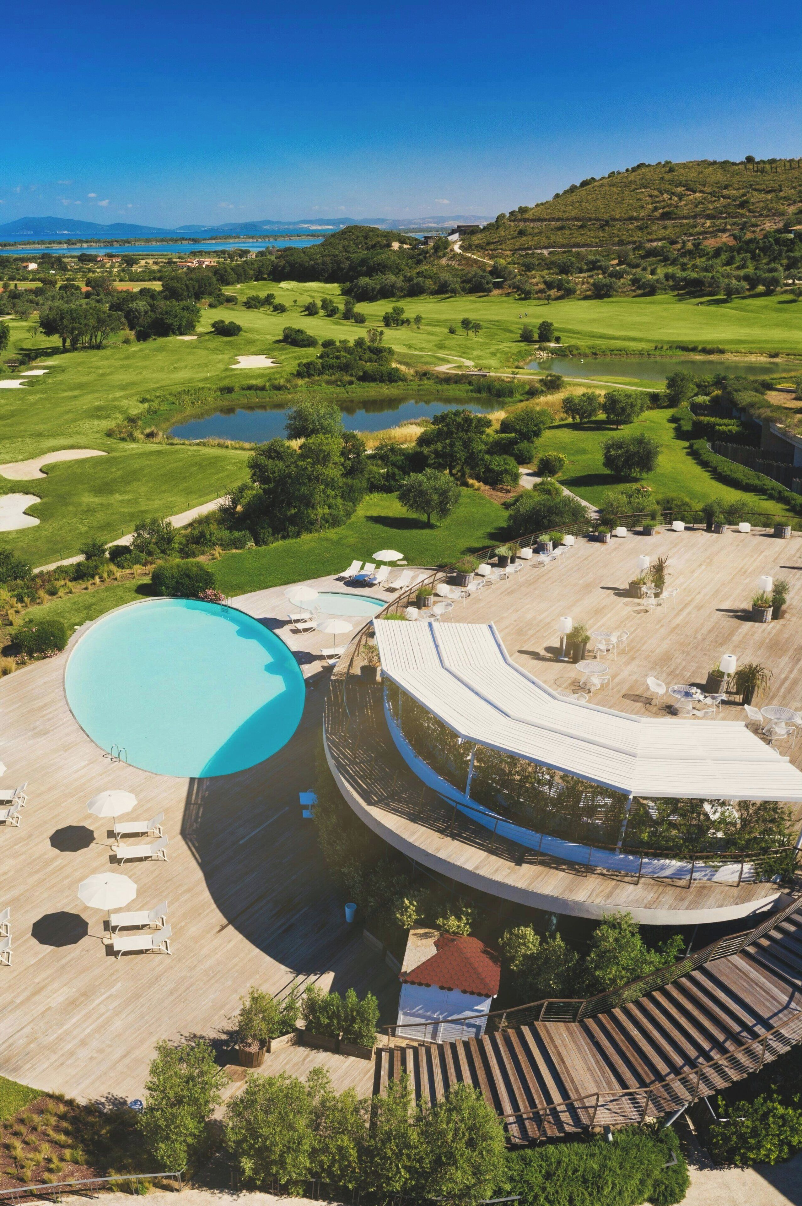 A resort with a large swimming pool, a covered outdoor area with seating, and a view of a golf course and hills in the background.
