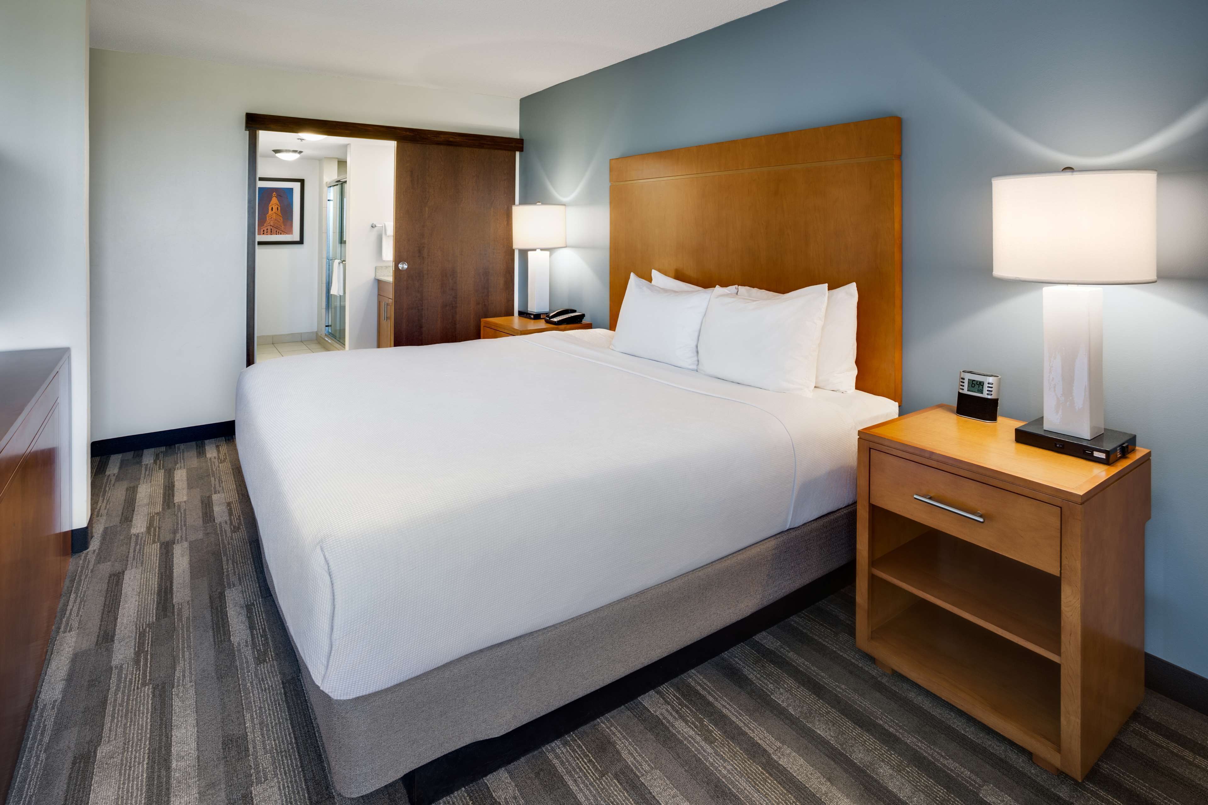 Habitación de hotel con una cama grande, cabecero de madera, lámparas de noche, una mesita de noche con teléfono y lámpara, y otra mesita de noche con cajón.