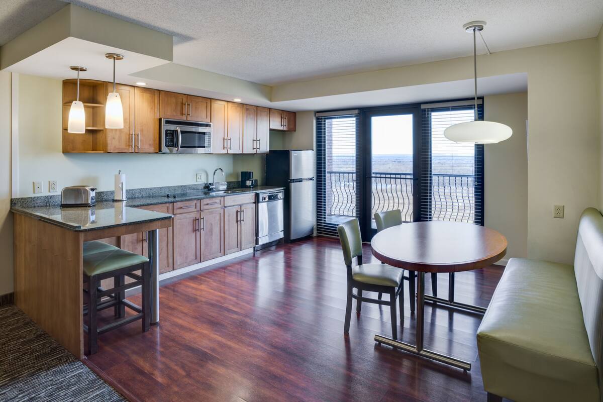 Una cocina moderna con armarios de madera, microondas y refrigerador. Hay un comedor con mesa y sillas, y un balcón con vistas.