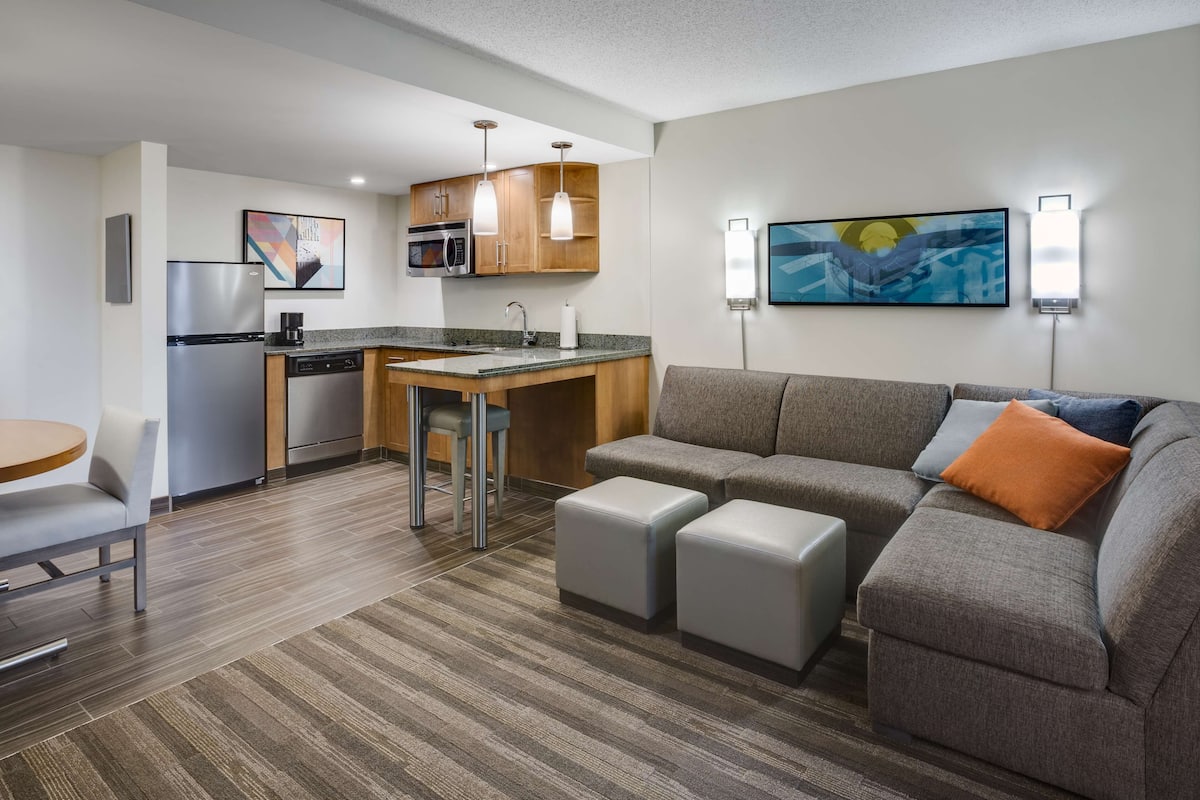 Una habitación de hotel moderna con kitchenette, un sofá gris con un cojín naranja y un comedor con una pequeña mesa y sillas.