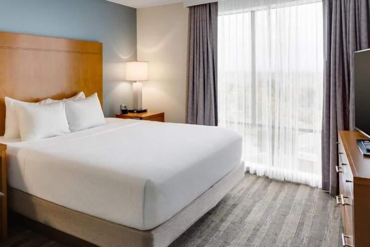 Habitación de hotel con una cama grande, un cabecero de madera, una mesita de noche con una lámpara, una silla y un escritorio con una computadora.