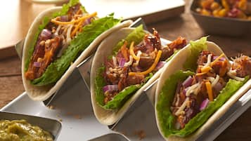 Trois tacos garnis de laitue, d’oignons rouges et de fromage râpé, présentés sur un plateau blanc, accompagnés d’un verre de boisson fraîche et de quartiers de citron vert.