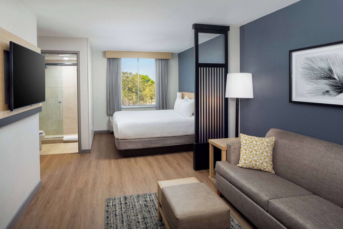 A hotel room with a bed, a sofa, a TV, a window with a view, and a painting on the wall.