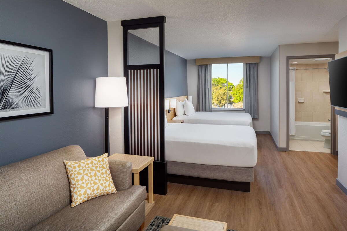 A hotel room with a sofa, two beds, a small table, a lamp, a mirror, and a view of the outdoors.