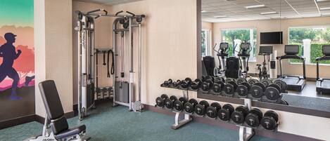 A gym with exercise equipment, a wall-mounted TV, and a window with a view of greenery.