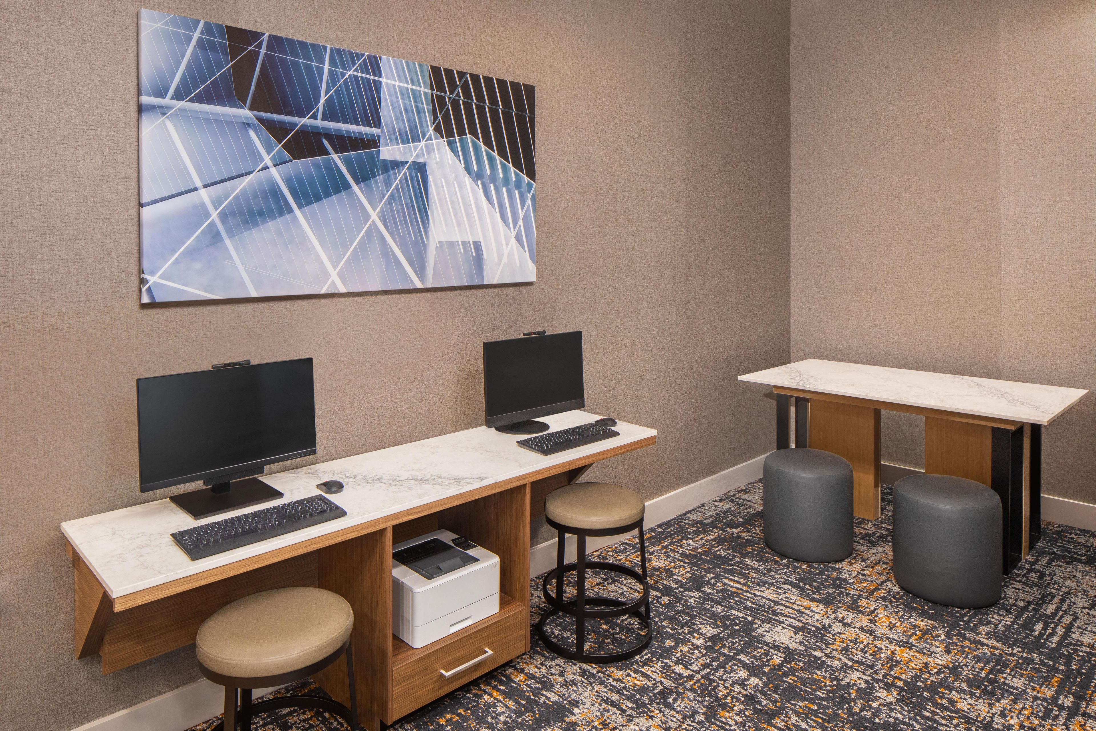 A modern office space with two desks, each with a computer monitor, keyboard, and a printer. There are two stools and a small table with a chair.