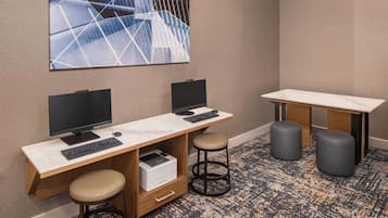 A modern office space with two desks, each with a computer monitor, keyboard, and a printer. There are two stools and a small table with a chair.