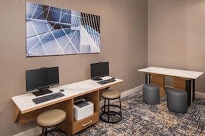 A modern office space with two desks, each with a computer monitor, keyboard, and a printer. There are two stools and a small table with a chair.