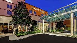 A Hyatt Place hotel entrance with a covered walkway, glass doors, and a landscaped area with trees and shrubs. - Hyatt Place Lake Mary/Orlando North (Lake Mary)