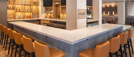 Zona de bar con una barra larga, varios taburetes y estantes con botellas.