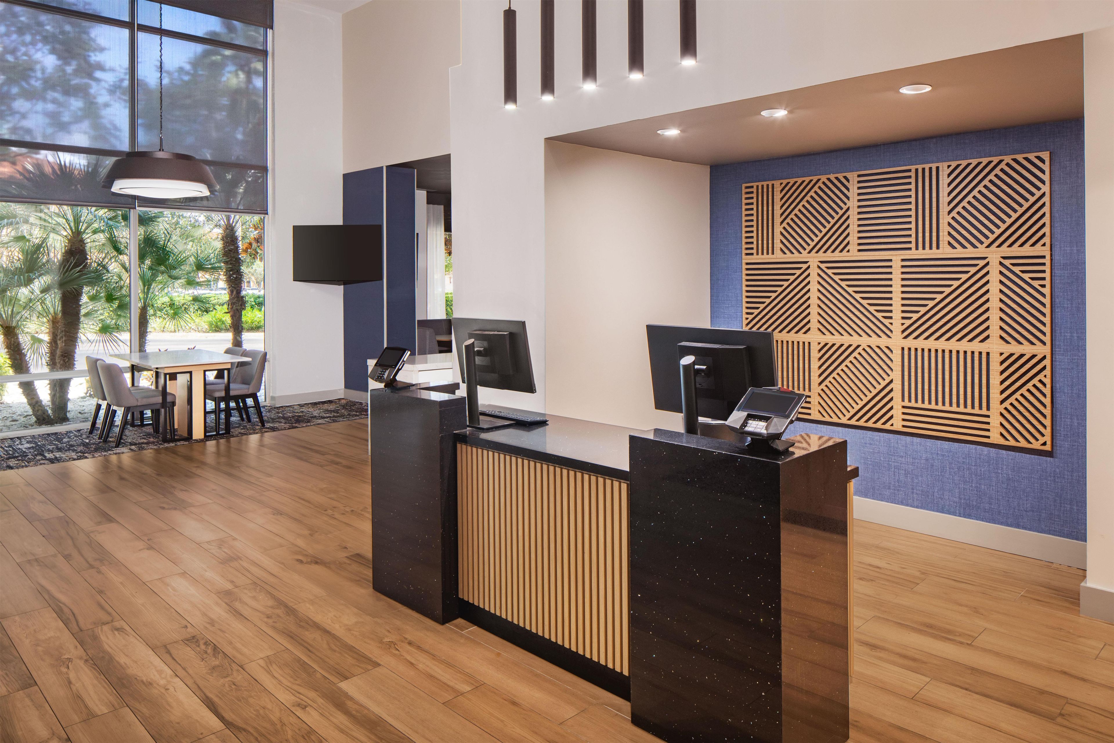 A modern reception desk with a flat-screen TV, a computer, and a phone. There is a seating area with chairs and a table, and a large window with a view of palm trees.