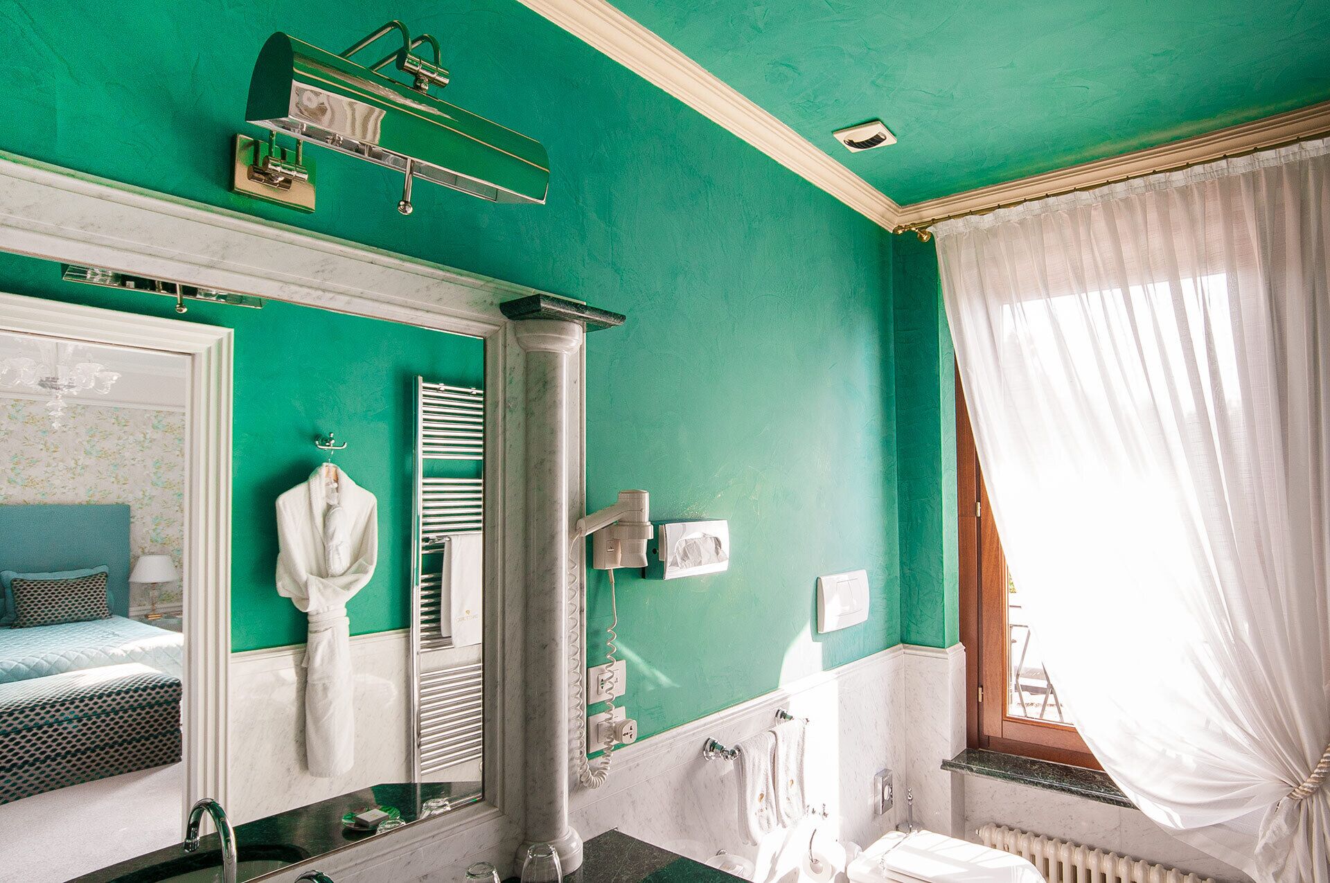 Un bagno con una parete verde, una vasca da bagno bianca, uno specchio, un porta asciugamani e una finestra con tende bianche.