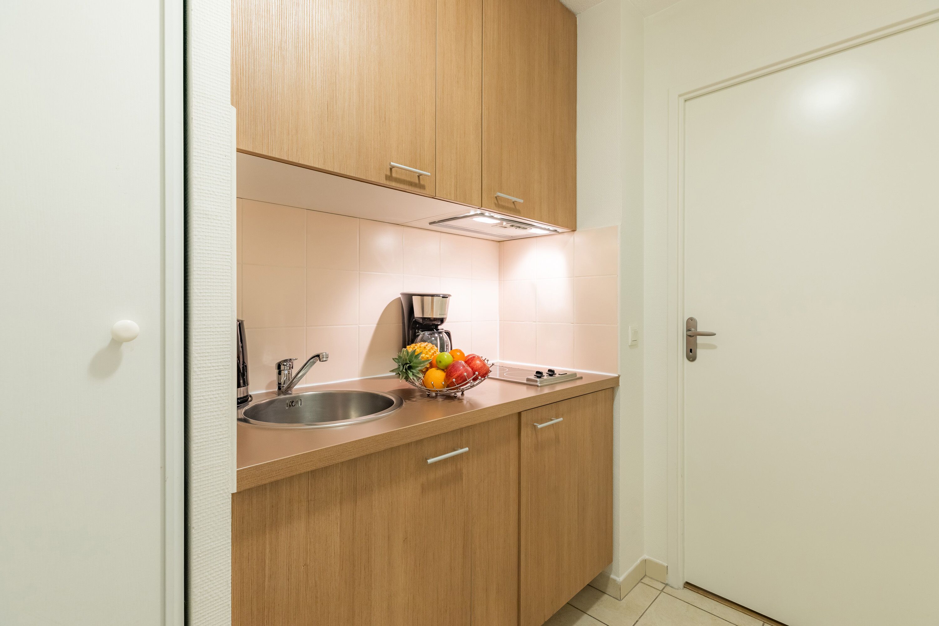 A compact kitchen with wooden cabinets, a stainless steel sink, and a fruit bowl on the countertop.