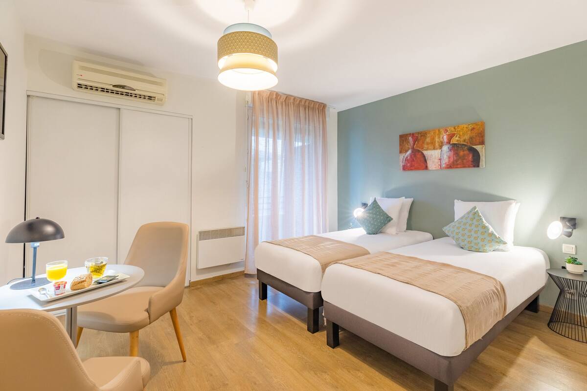 A hotel room with two beds, a dining area with a table and chairs, a ceiling fan, and a painting on the wall.