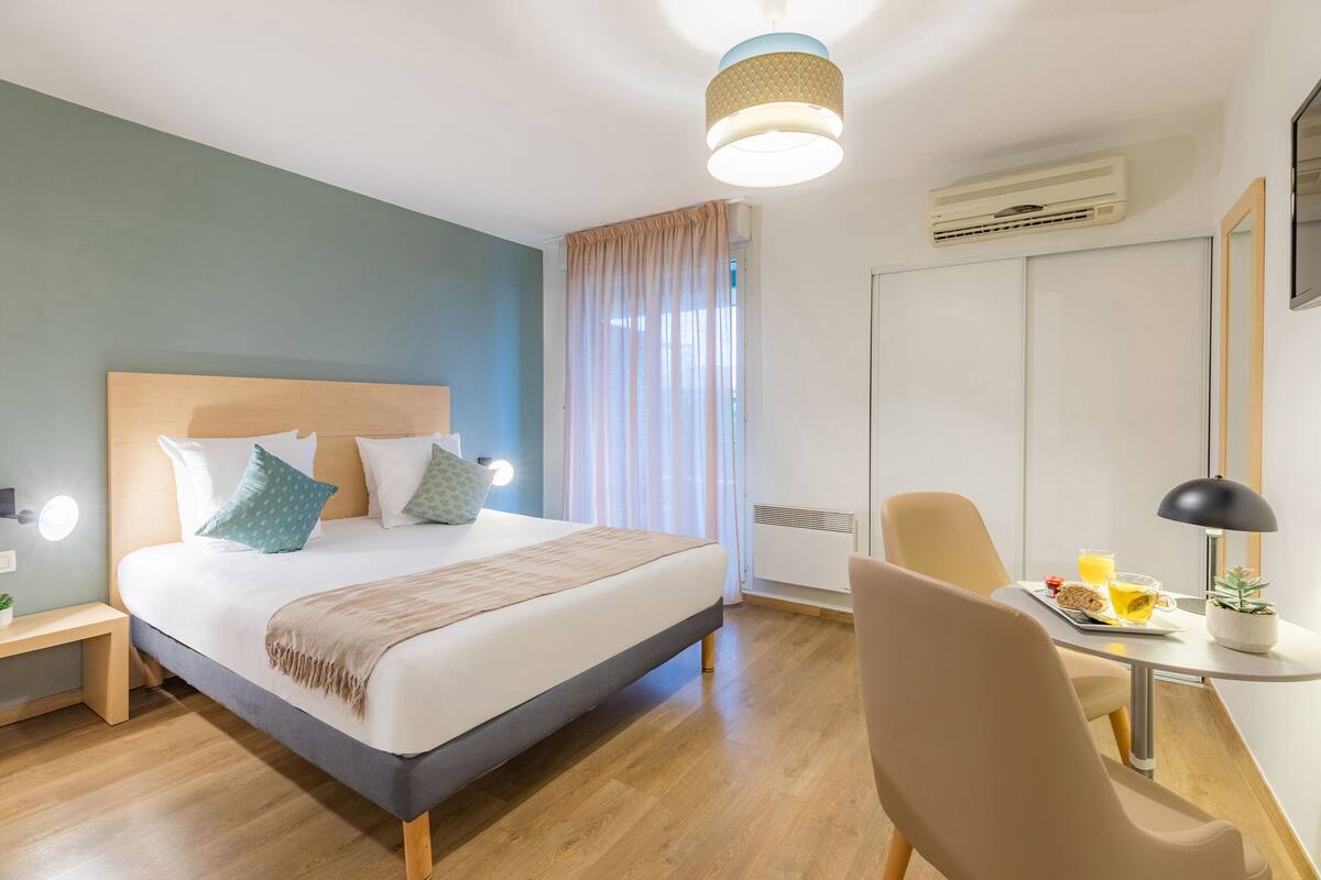 A modern hotel room with a large bed, bedside table, and a desk with a chair.