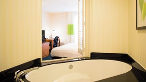 Casa de banho de hotel com uma banheira grande, um espelho refletindo um quarto com cama, secretária e cadeira.