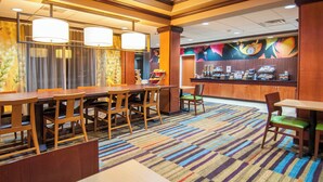 Uma área de refeições de hotel com mesas e cadeiras de madeira, um tapete listrado e vibrante, e um balcão com diversos alimentos e uma máquina de café.