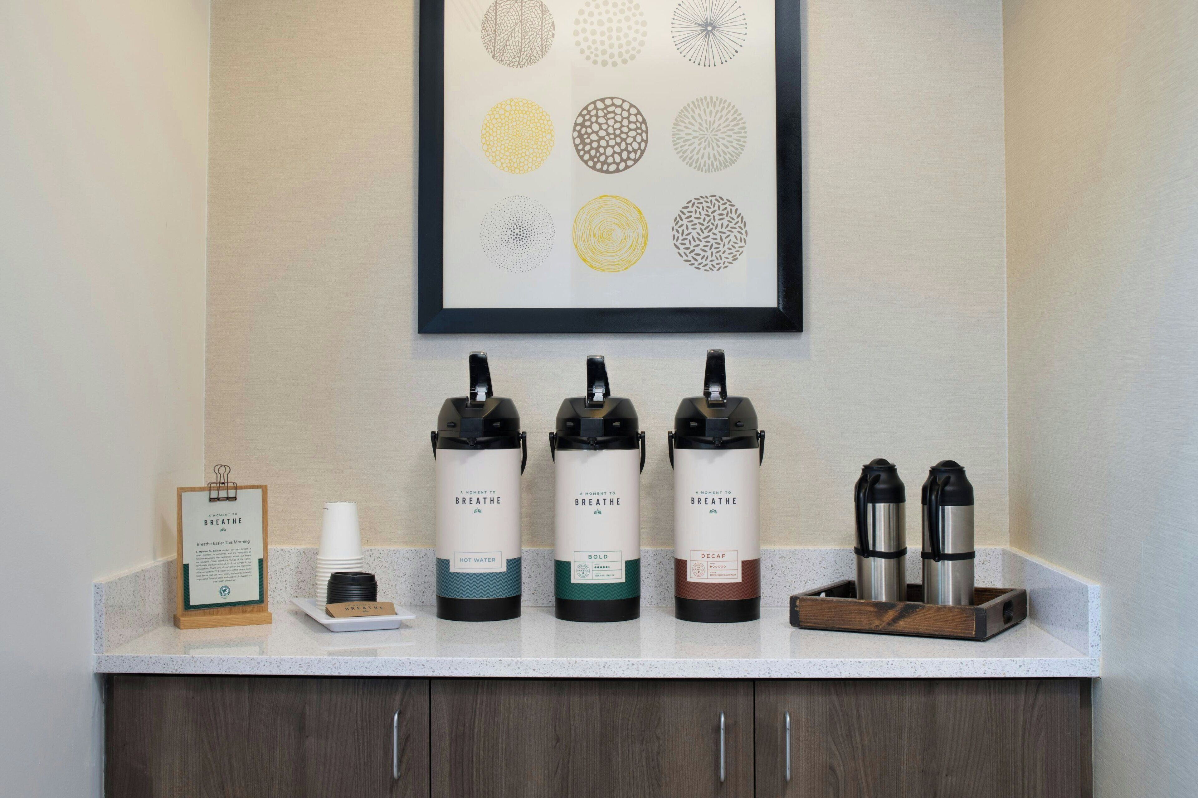 A countertop with water dispensers labeled "BREATHE," "HOT WATER," "BOLD," and "DECAF," along with a framed artwork above.