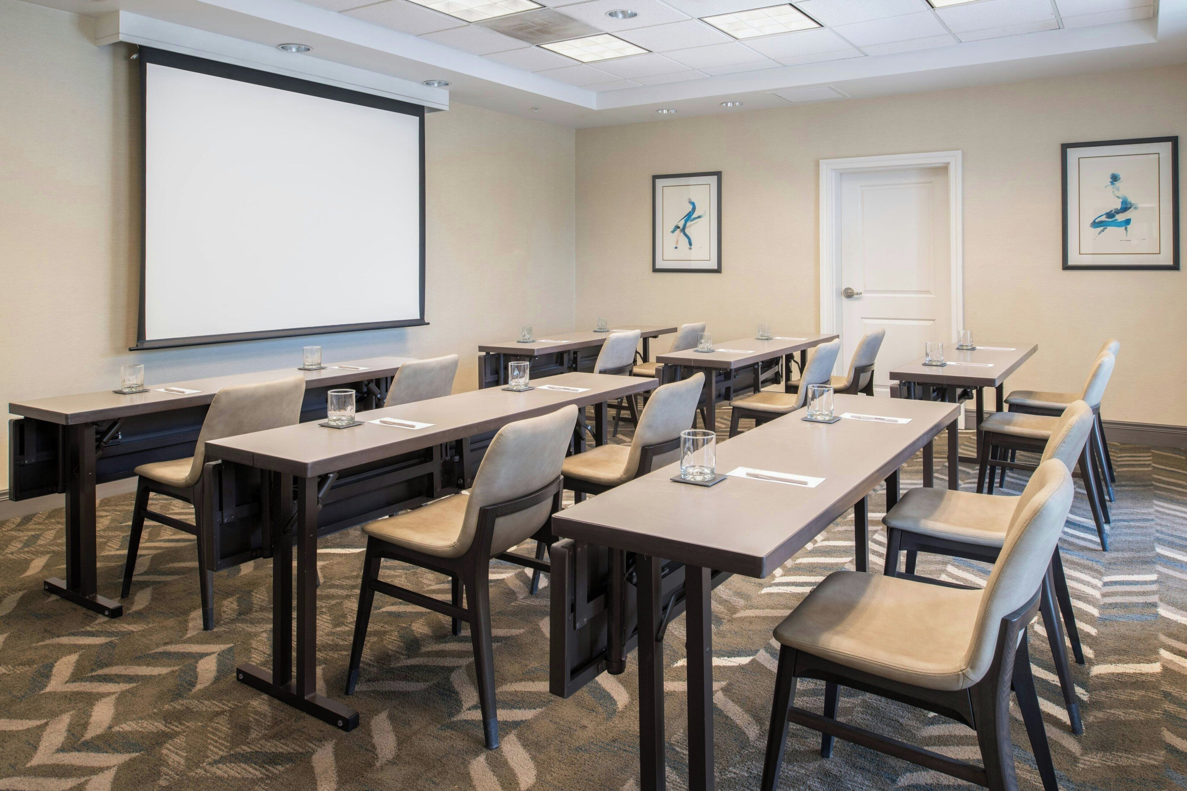 A conference room with a large projection screen, multiple tables with chairs, and framed artwork on the walls.