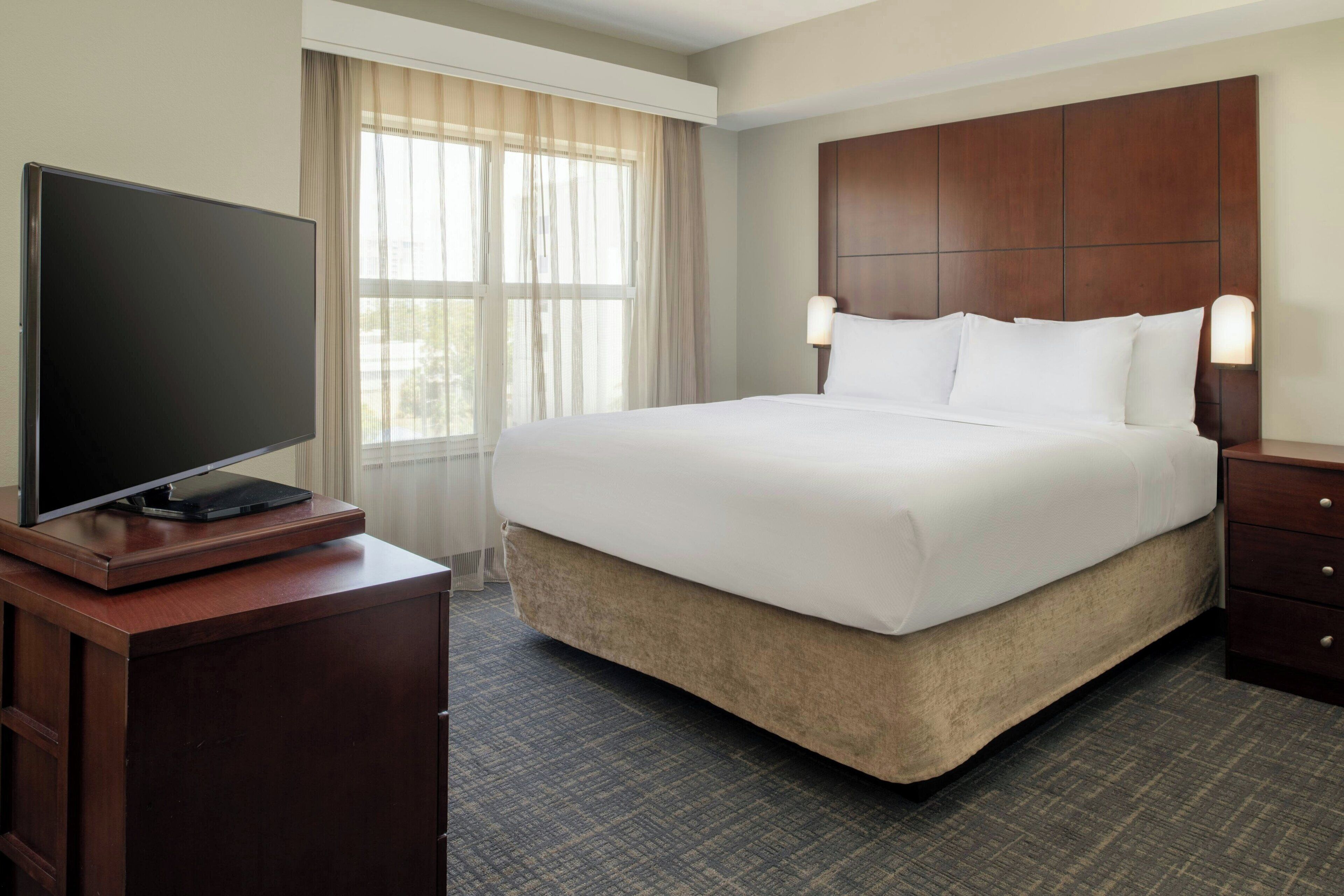 Habitación de hotel con una cama grande, un televisor de pantalla plana sobre un aparador de madera y una ventana con cortinas translúcidas.