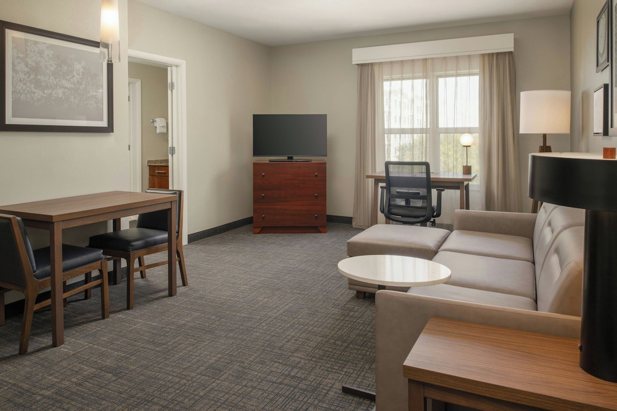 Habitación de hotel con un escritorio de madera, dos sillas, un sofá, un televisor, una lámpara y una ventana con cortinas.
