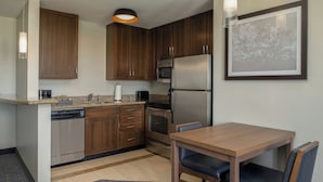A compact kitchen with wooden cabinets, a stainless steel refrigerator, a microwave, and a dining area with a table and chairs. - Residence Inn by Marriott Clearwater Downtown (Clearwater)