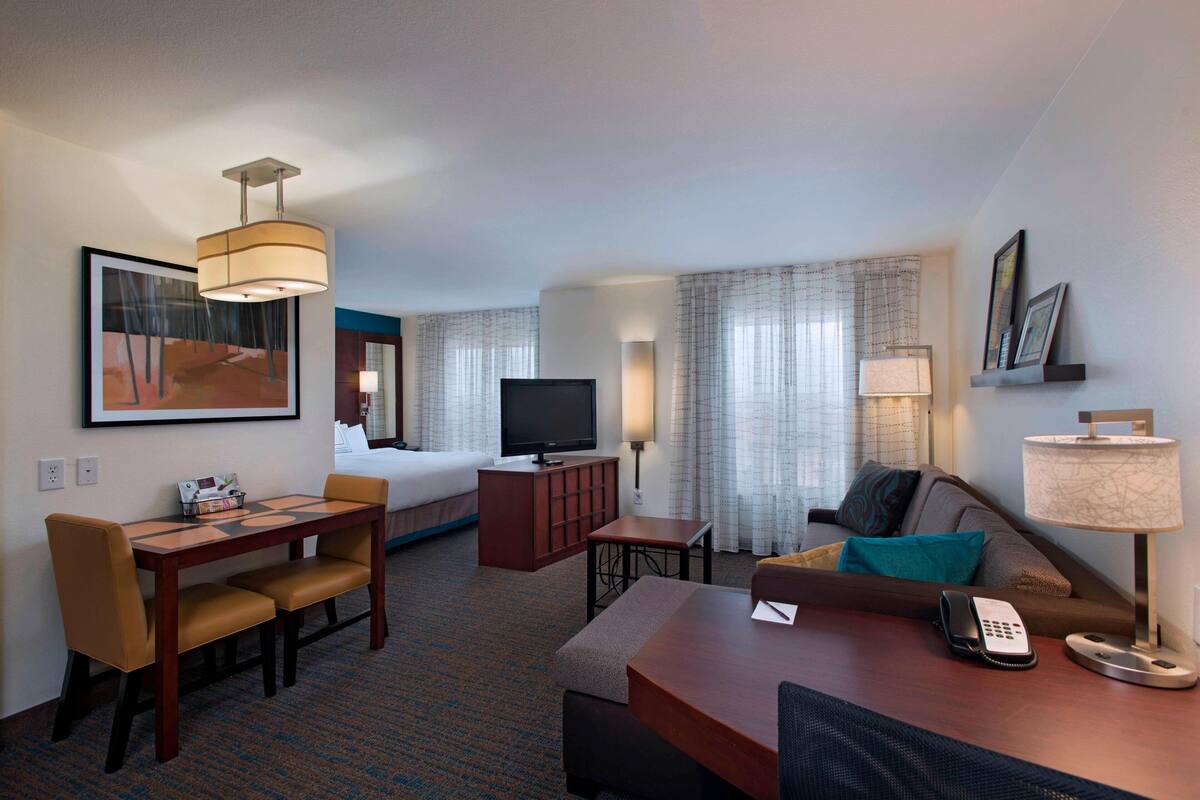 A hotel room with a bed, a TV, a desk with a telephone, a lamp, and a painting on the wall.