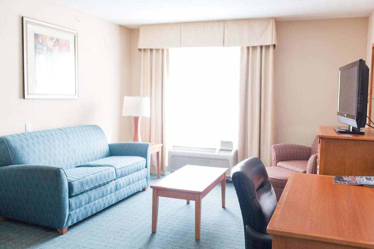 Ein Hotelzimmer mit einem blauen Sofa, einem Holztisch, einem schwarzen Sessel, einem Fernseher und einem Fenster mit Vorhängen.