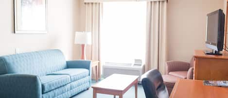 Habitación de hotel con un sofá azul, una mesa de madera, una silla negra, un televisor y una ventana con cortinas.