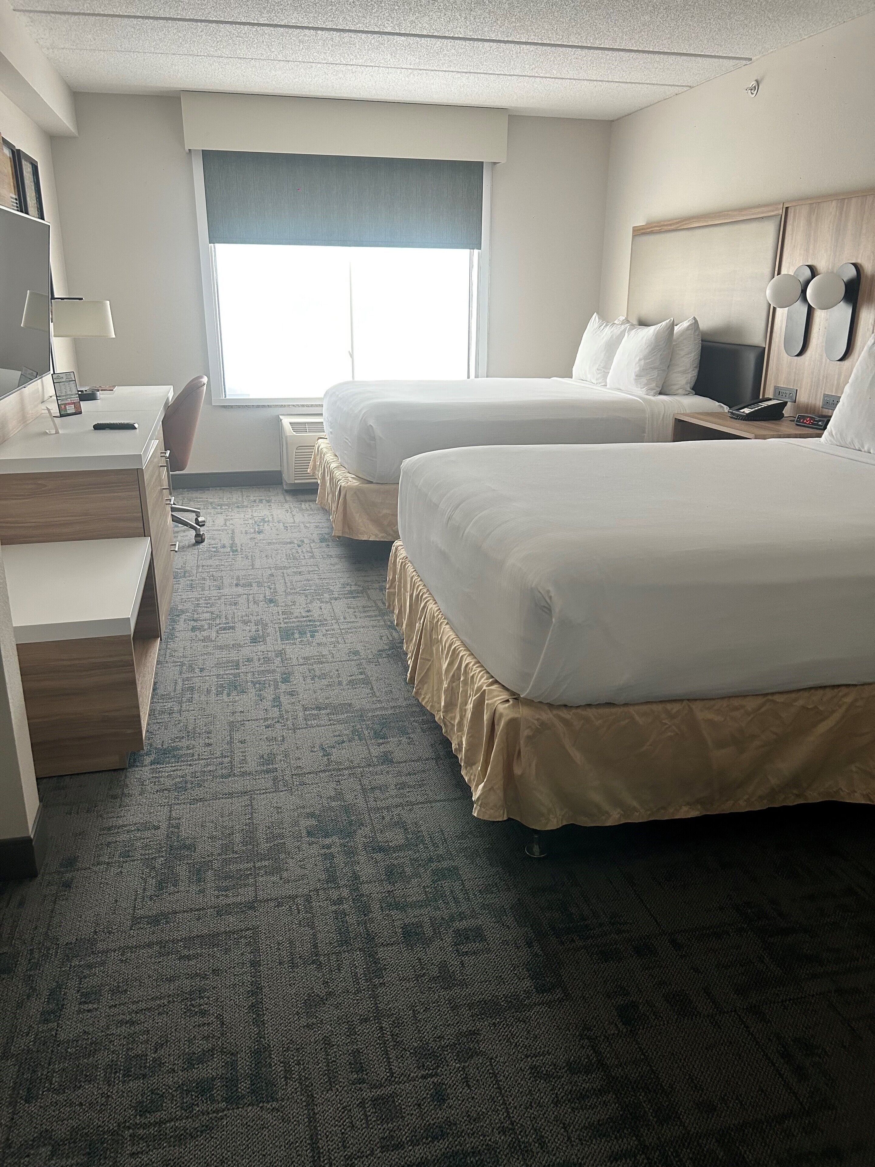 A hotel room with two beds, a desk, a chair, a lamp, and a window.