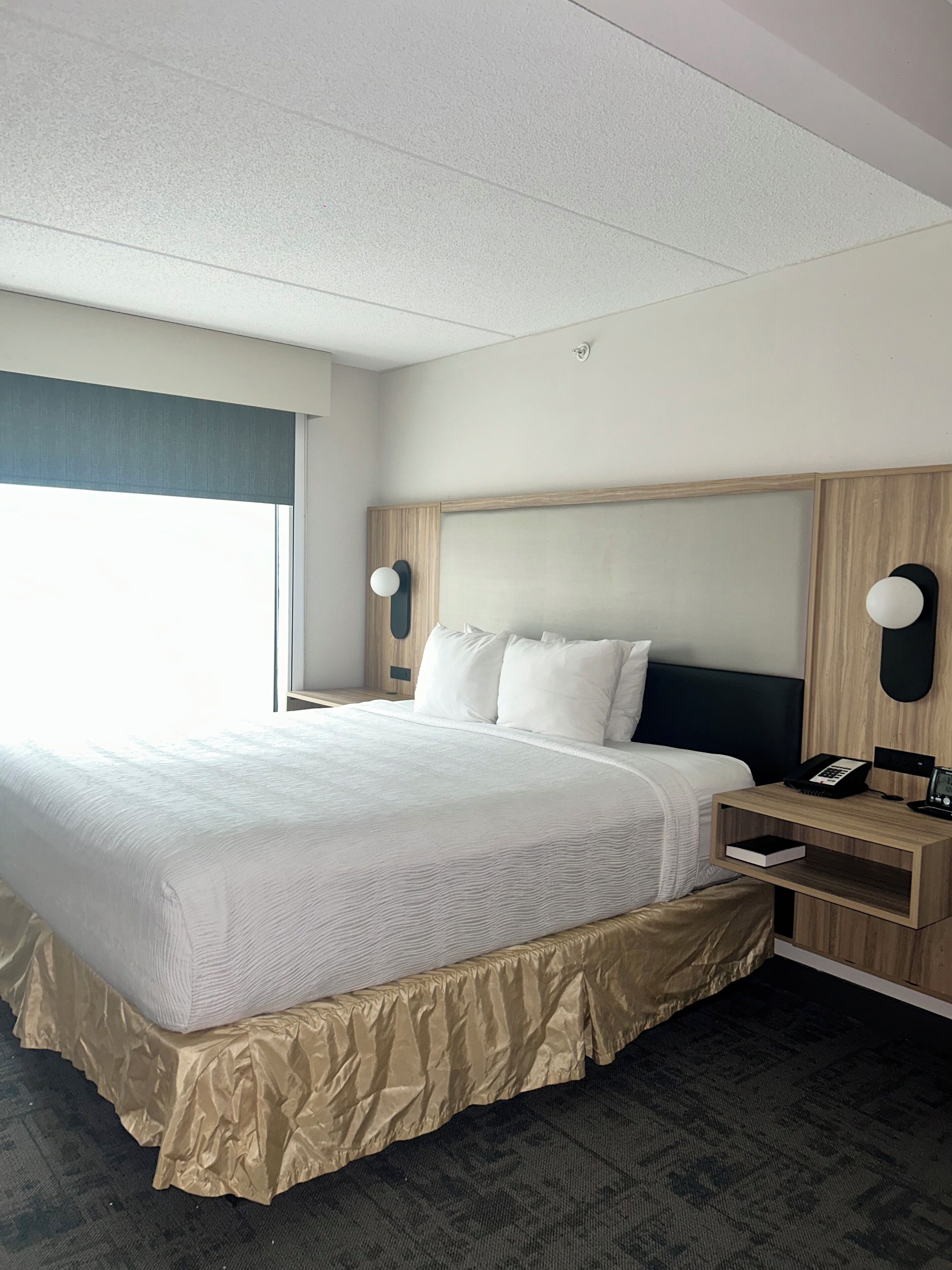 A hotel room with a large bed, a wooden headboard, a bedside table with a phone, and a window with a view.