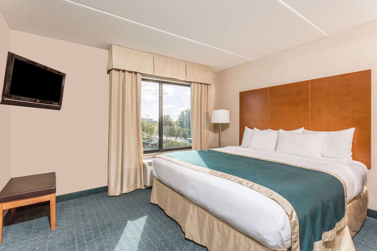 Ein Hotelzimmer mit Bett, einem an der Wand montierten Fernseher, einem Fenster mit Vorhängen und einem Nachttisch.