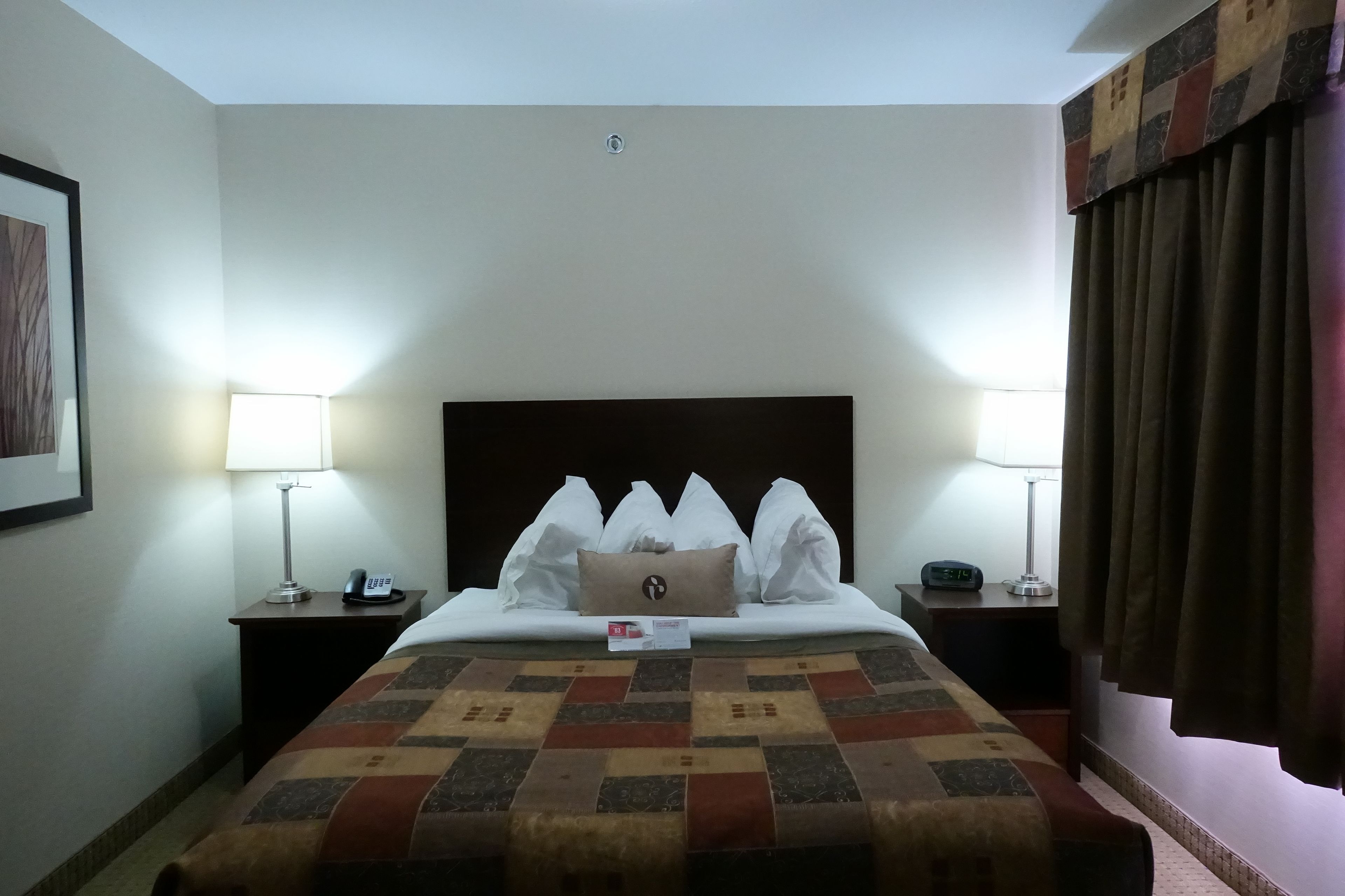 A hotel room with a large bed, two bedside tables, a nightstand, a lamp, a clock, and a curtain.