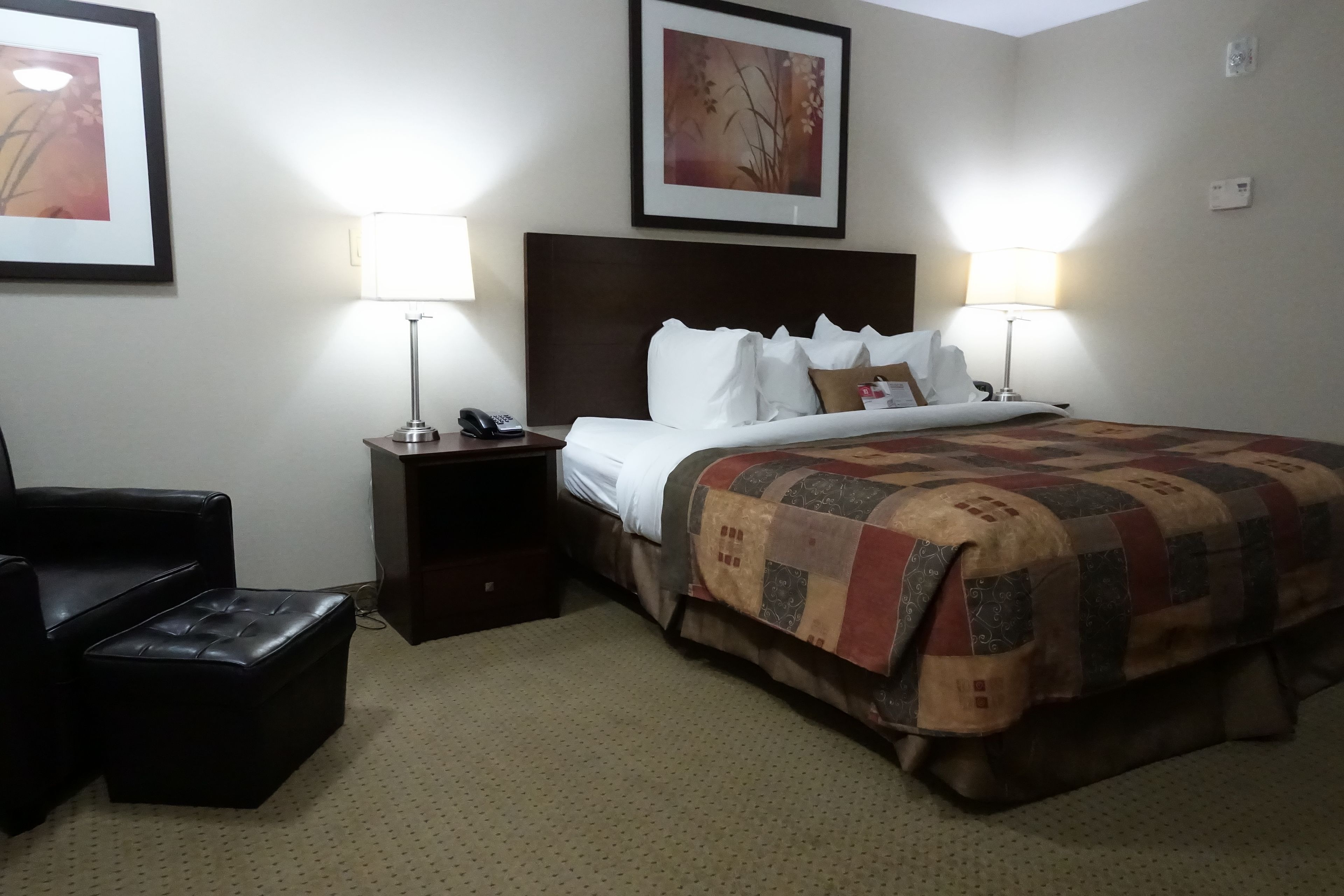 A hotel room with a bed, two bedside lamps, a nightstand with a phone, a black leather chair, and a small ottoman.