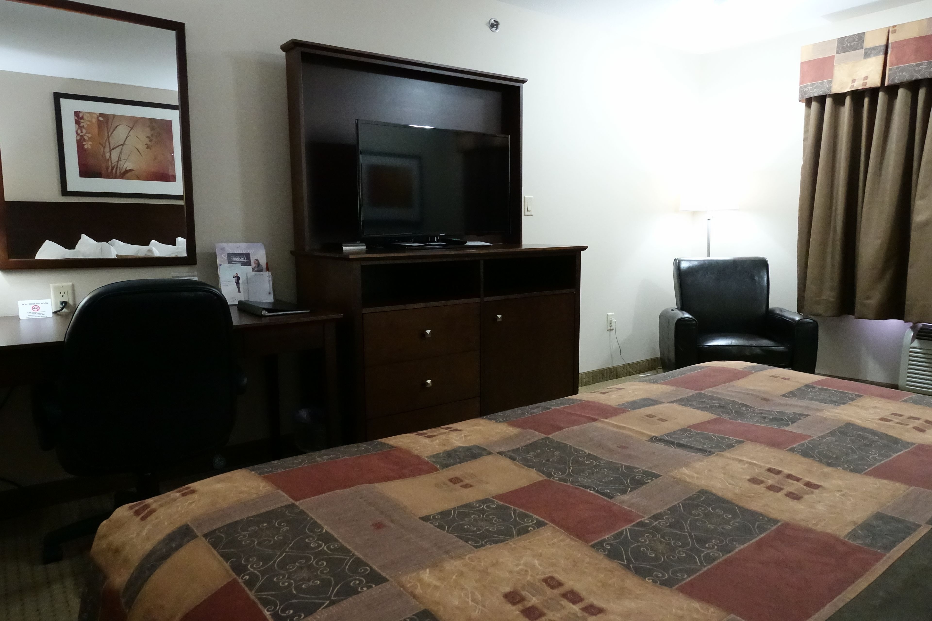 Habitación de hotel con una cama grande, un escritorio con computadora, un televisor sobre un mueble de madera, una silla de oficina negra y una ventana con cortinas.